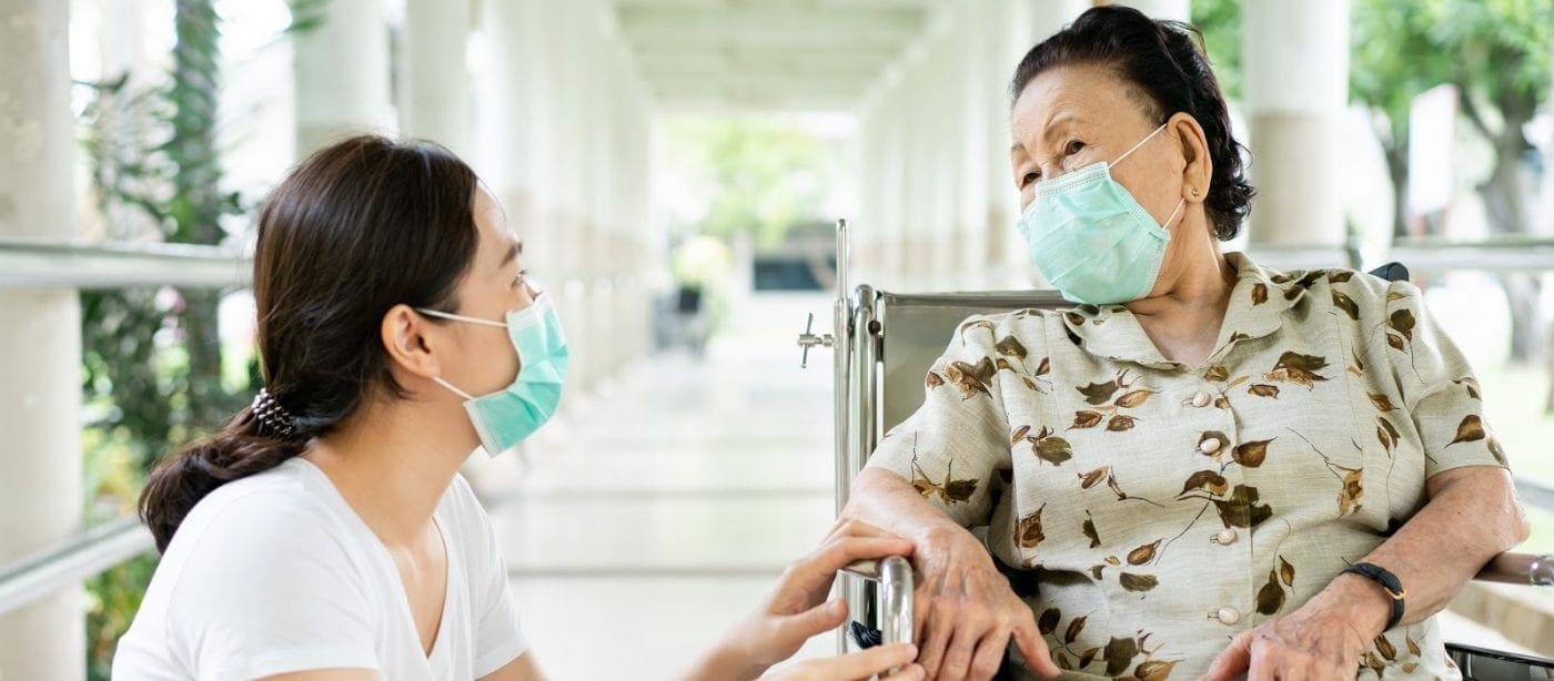 Younger adult speaking with an older adult in a wheelchair in a senior living facility corridor, both wearing face masks.