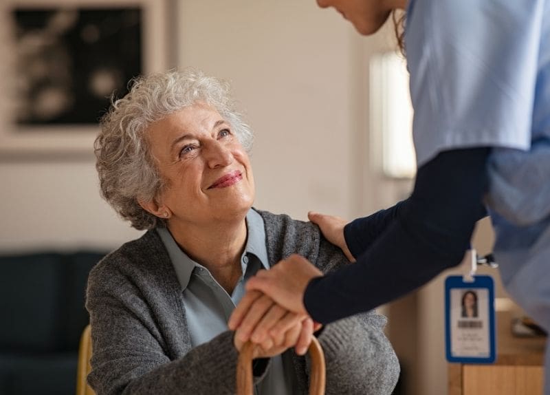 Elderly woman holding a cane smiling at a caregiver who gently holds her hands indoors.