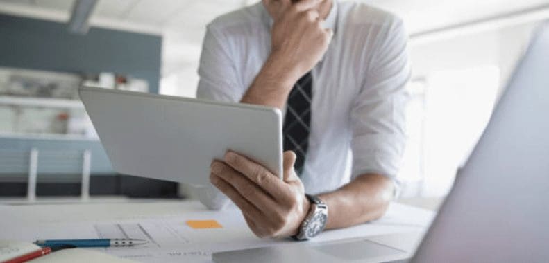 Person in business attire holding a tablet and standing at a desk with a laptop and papers.