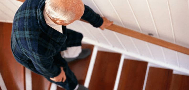 An elderly resident descending a wooden staircase indoors while holding the handrail for support.