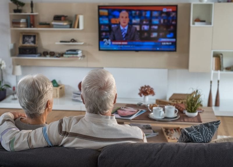 Two seniors seated on a couch watching news on a television in a comfortable living room with a coffee table and decor.