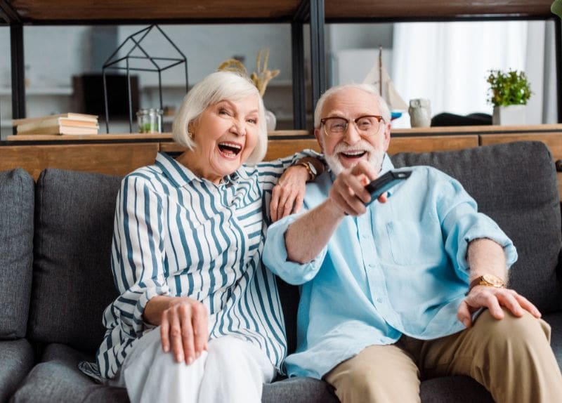 Two senior residents sitting on a couch smiling and using a remote control in a cozy living room setting.