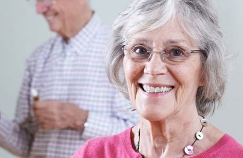 Smiling senior woman wearing glasses indoors with a senior man blurred in the background.