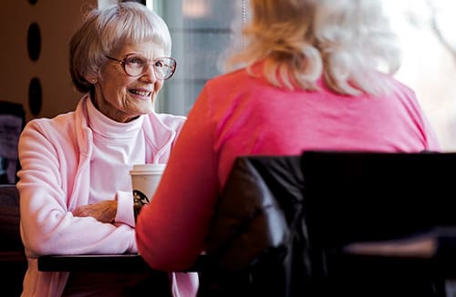 Two seniors sitting at a table indoors, having a conversation over coffee.