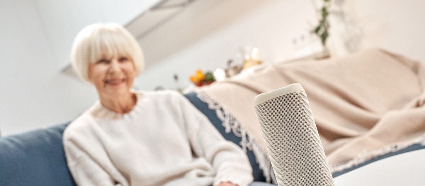 Elderly woman sitting on a couch with a smart speaker in a senior living community living room.