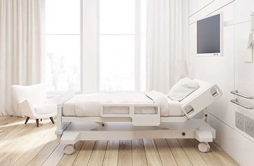Bright senior living care room with adjustable bed, white armchair, and wall-mounted television near large windows with curtains.