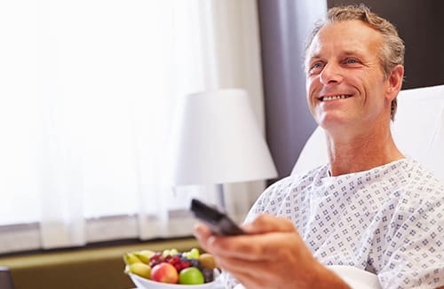 Man in a senior living room holding a remote control and smiling with a bowl of fresh fruit nearby.