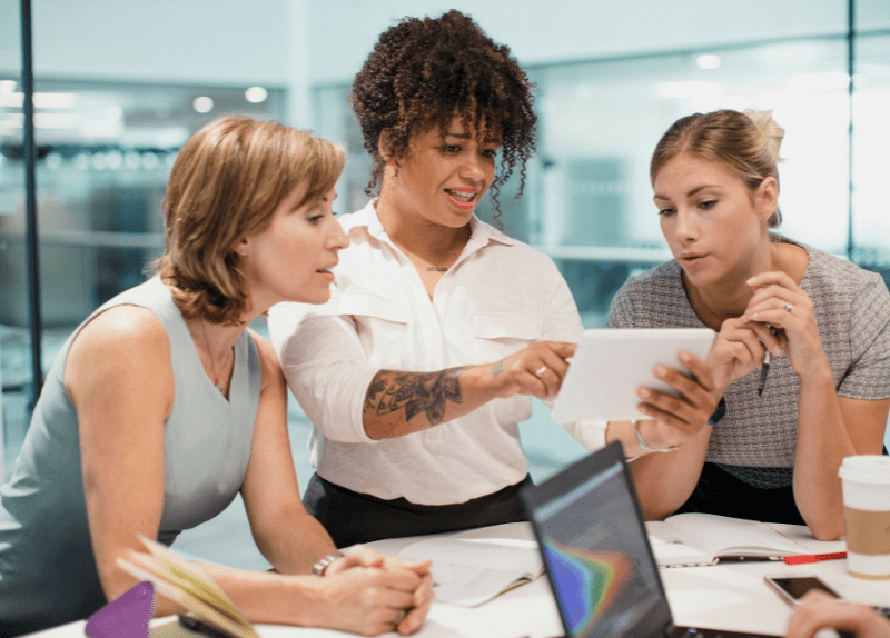 Three professional women collaborating around a table reviewing content on a tablet in an office meeting.
