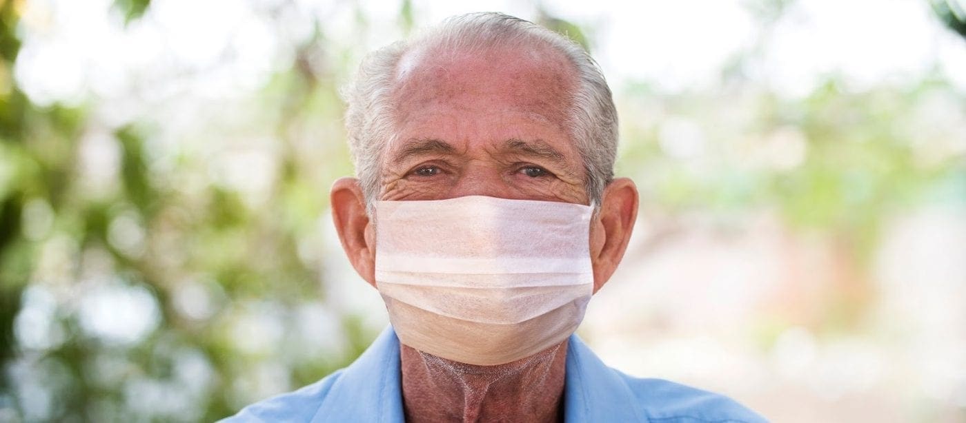 Elderly man wearing a protective face mask outdoors with greenery in the background.