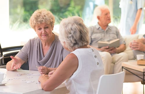 Two senior women working together on a puzzle at a table in a light-filled community room with other seniors visible in the background.