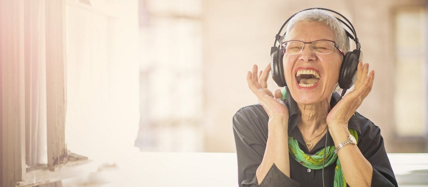 Elderly person enjoying music with headphones indoors near a window, expressing happiness and relaxation.