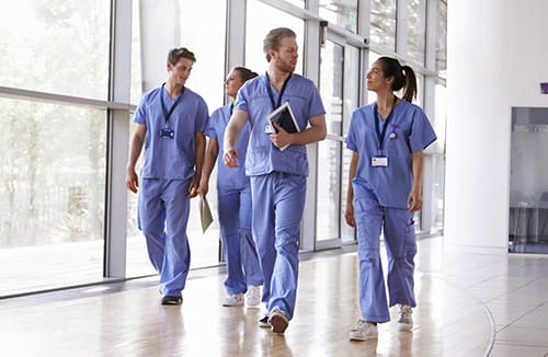 Four healthcare professionals in blue scrubs walking and conversing in a bright corridor with large windows.