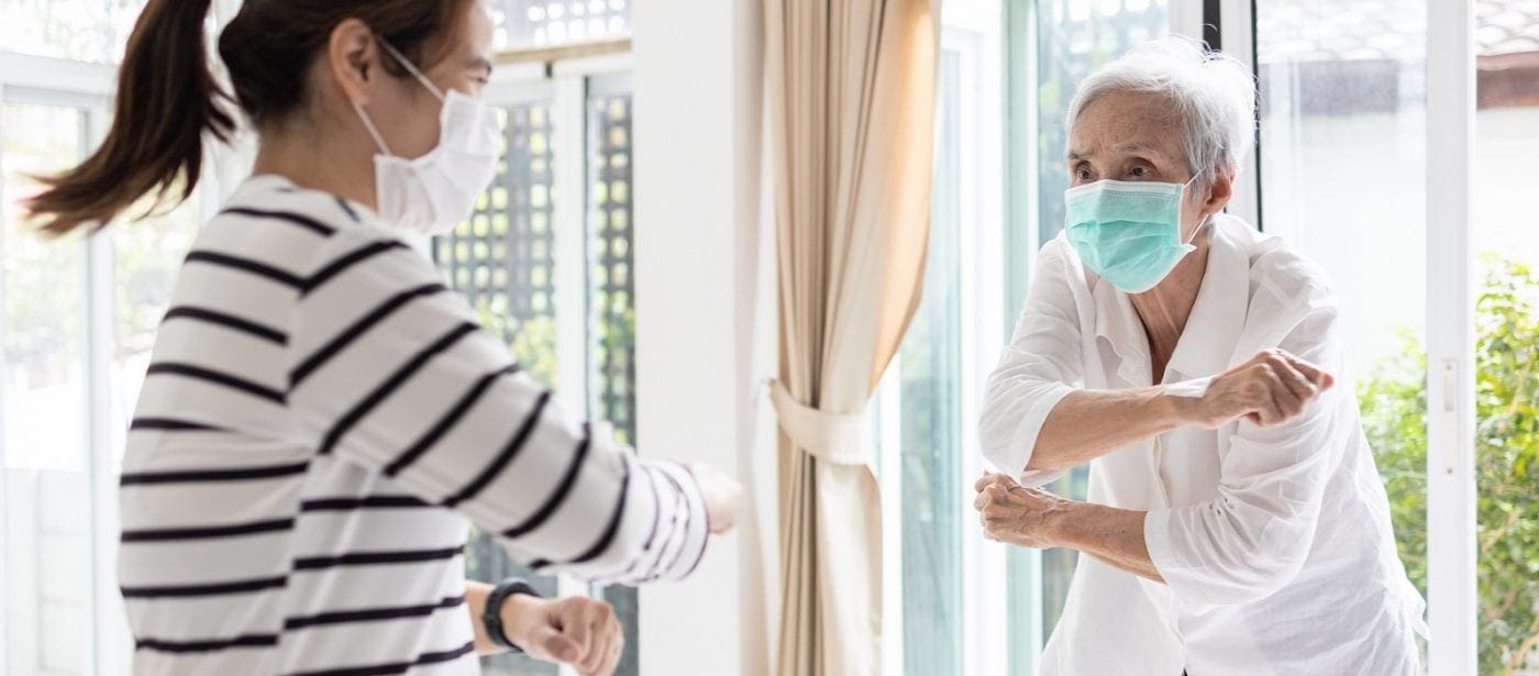 Two people wearing face masks exchanging an elbow greeting by a window inside a senior living community space.