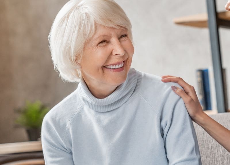 A senior woman smiling warmly while receiving a supportive hand on her shoulder in a comfortable community setting.