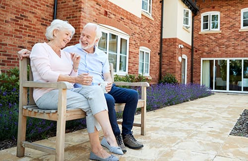 Two elderly residents sitting and chatting on a wooden bench outside a senior living community with brick walls and flowers.