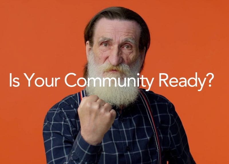 Elderly man with white beard wearing a blue checkered shirt and suspenders, posing with a raised fist in front of an orange background with a community readiness question.