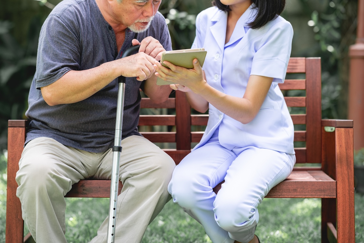 Elderly man with cane sitting on a bench outdoors while a caregiver shows him a tablet device.