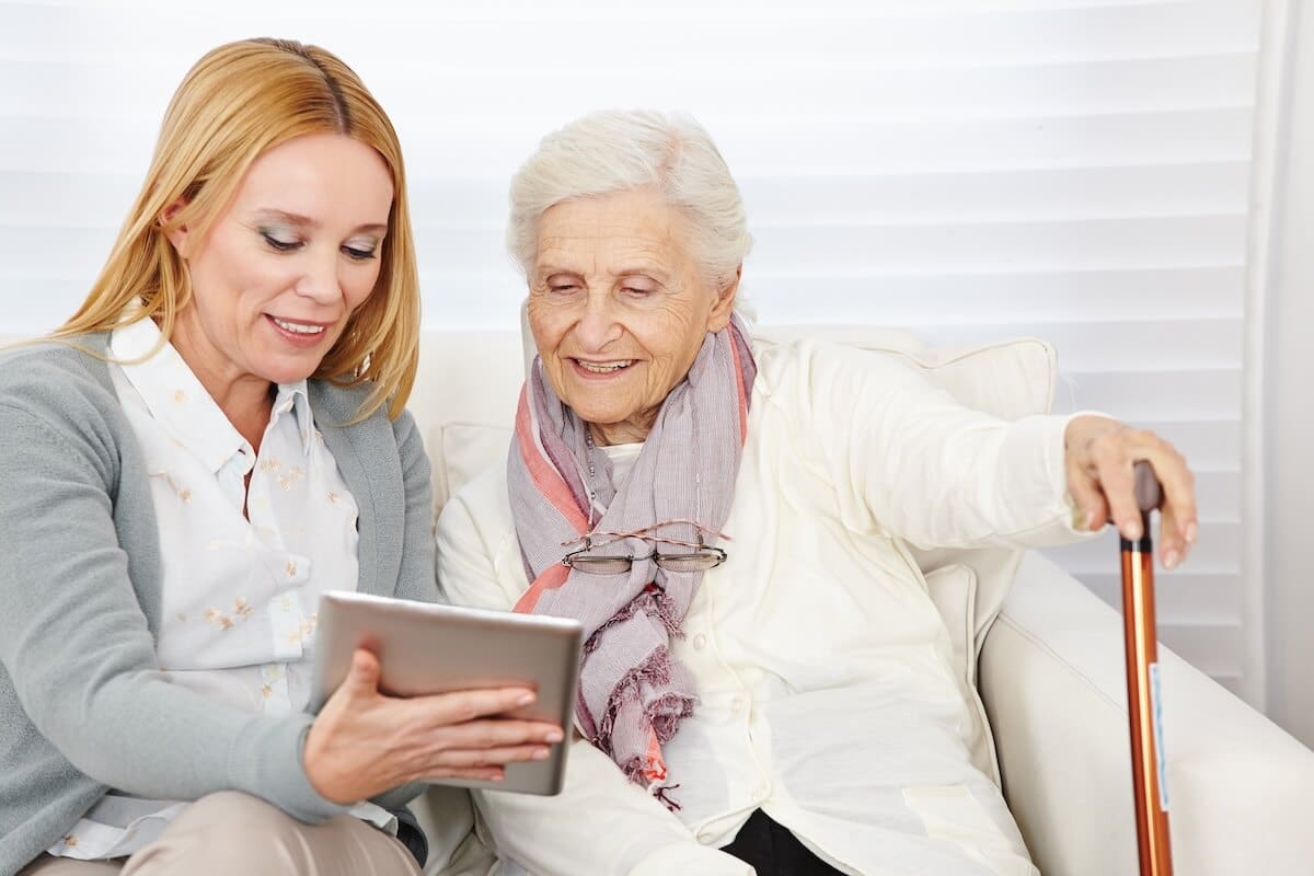 An elderly woman with a walking cane sits on a sofa with a younger woman who is showing her a digital tablet indoors.