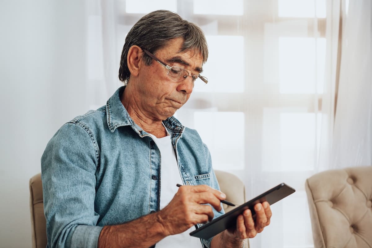 An older adult man sitting on a cushioned chair using a stylus to interact with a tablet in a bright room.