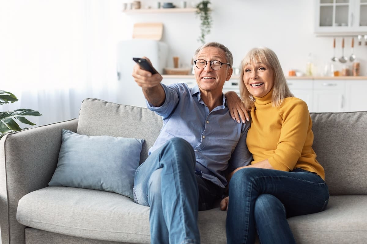 Older couple sitting on a couch smiling while holding a remote control in a bright living room.