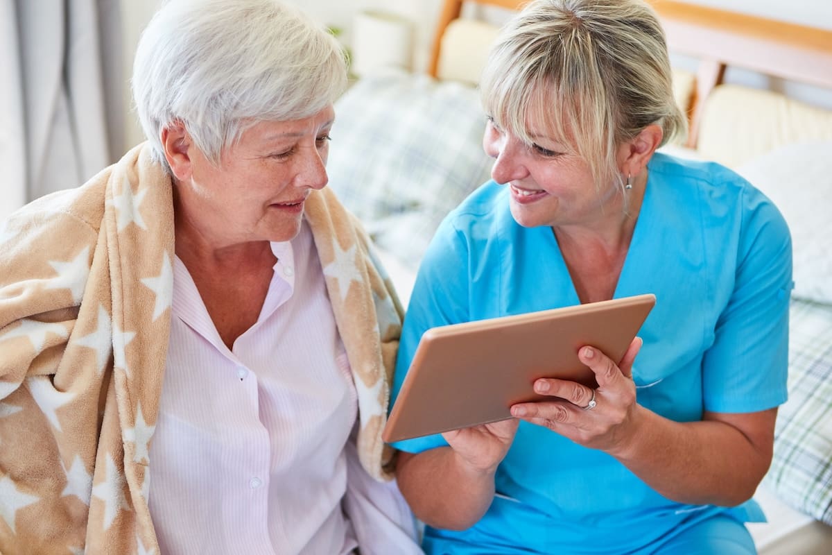 Healthcare professional in blue scrubs showing a tablet to an older woman wrapped in a blanket in a comfortable living area.