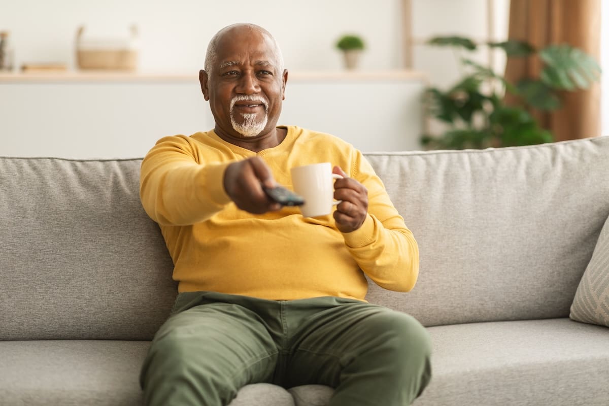 An older adult sitting on a couch holding a mug and pointing a remote control in a living room setting.