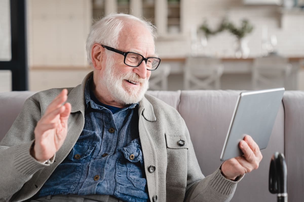 Elderly man sitting on a sofa waving while holding a tablet during a video call indoors.