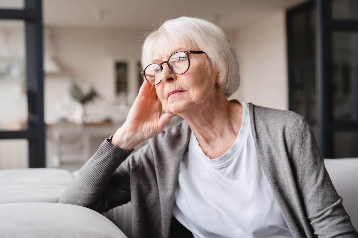 Elderly woman sitting on a couch indoors resting her head on her hand with a thoughtful expression.