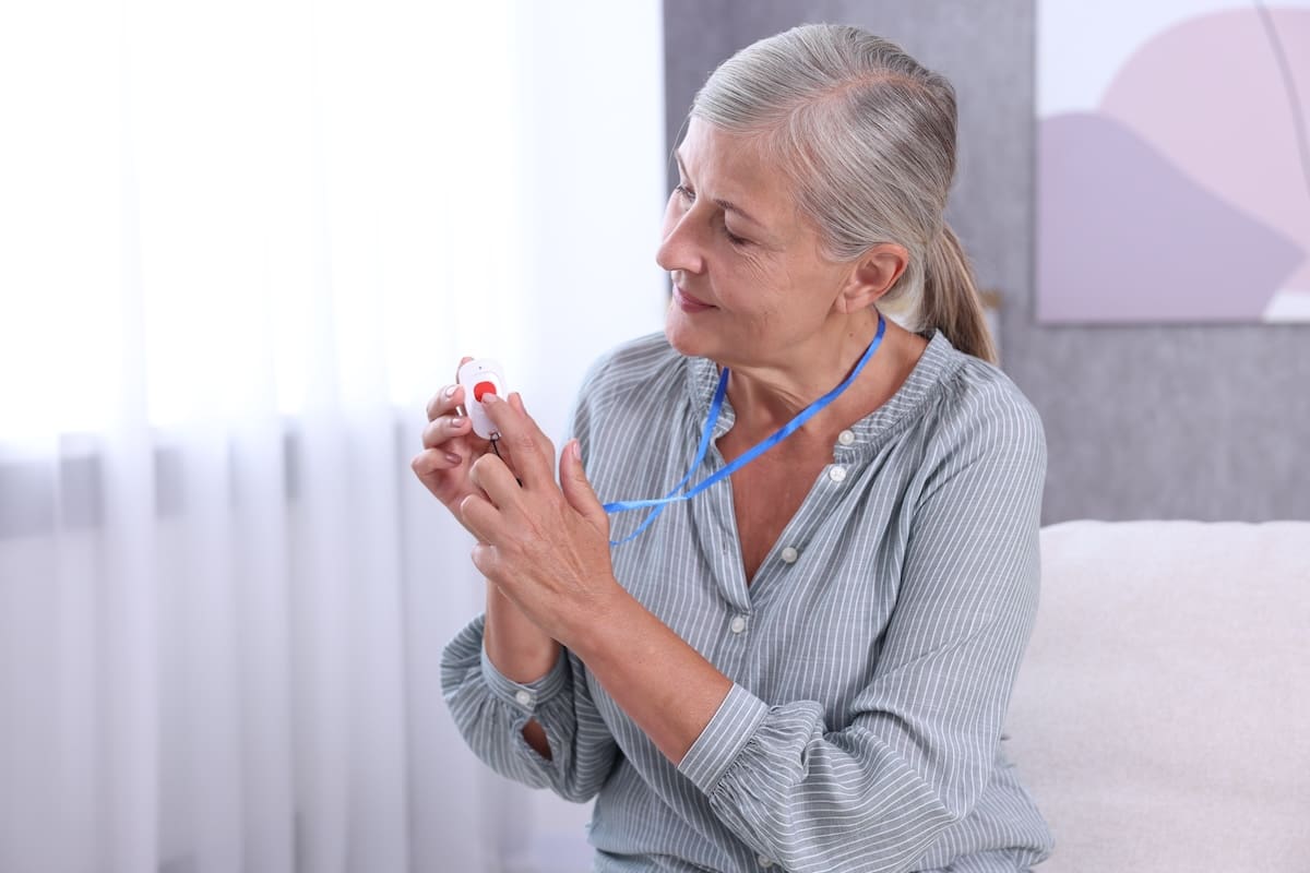 Older woman sitting and looking at a personal emergency alert device with blue lanyard in a bright room.