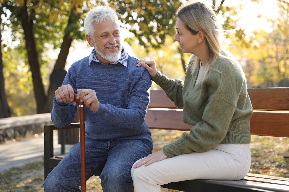 Two people sitting on a bench in a park surrounded by autumn trees, engaged in conversation with a walking cane resting beside them.