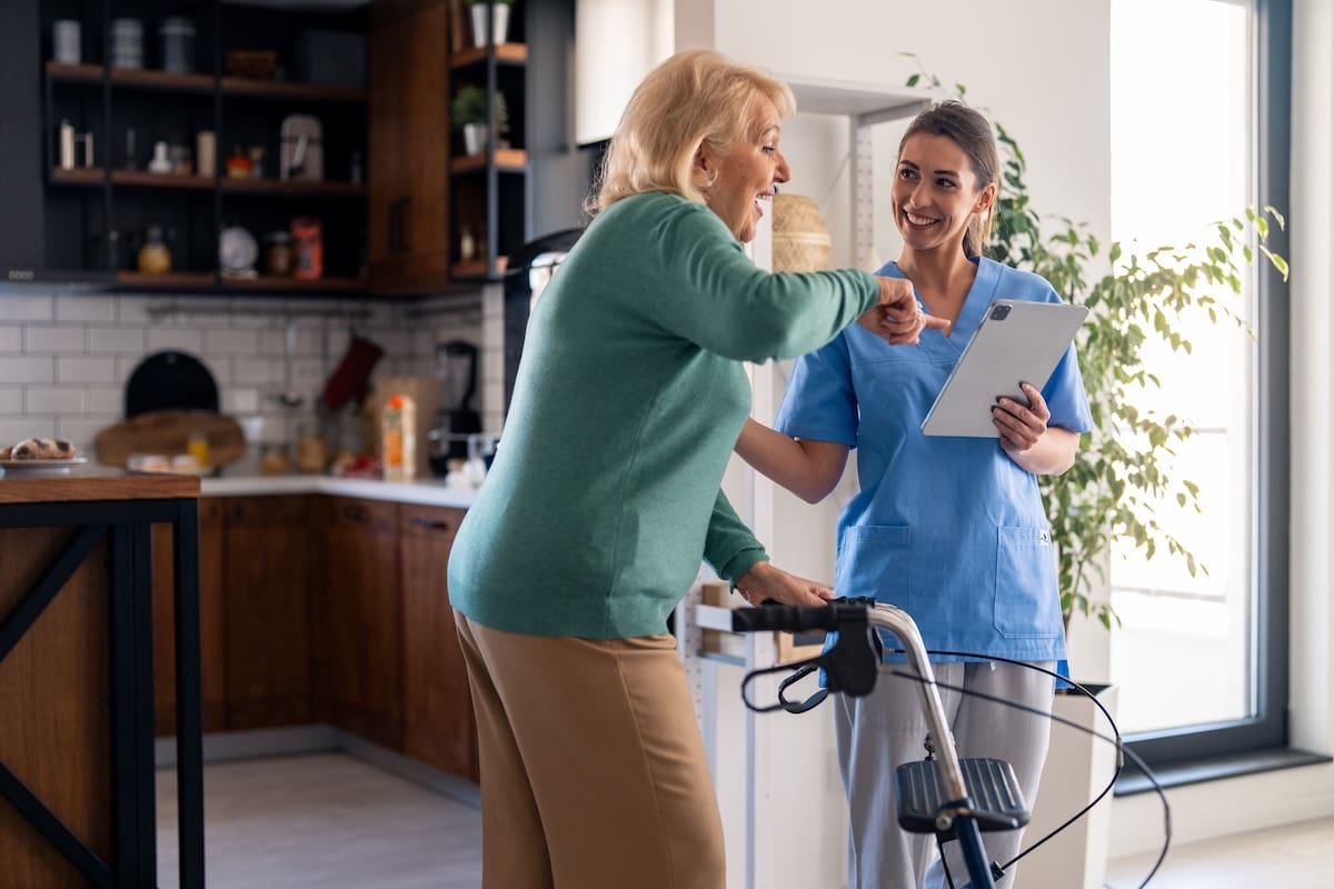 Healthcare worker engaging with a senior resident using a rollator walker in a kitchen setting while reviewing a digital tablet.