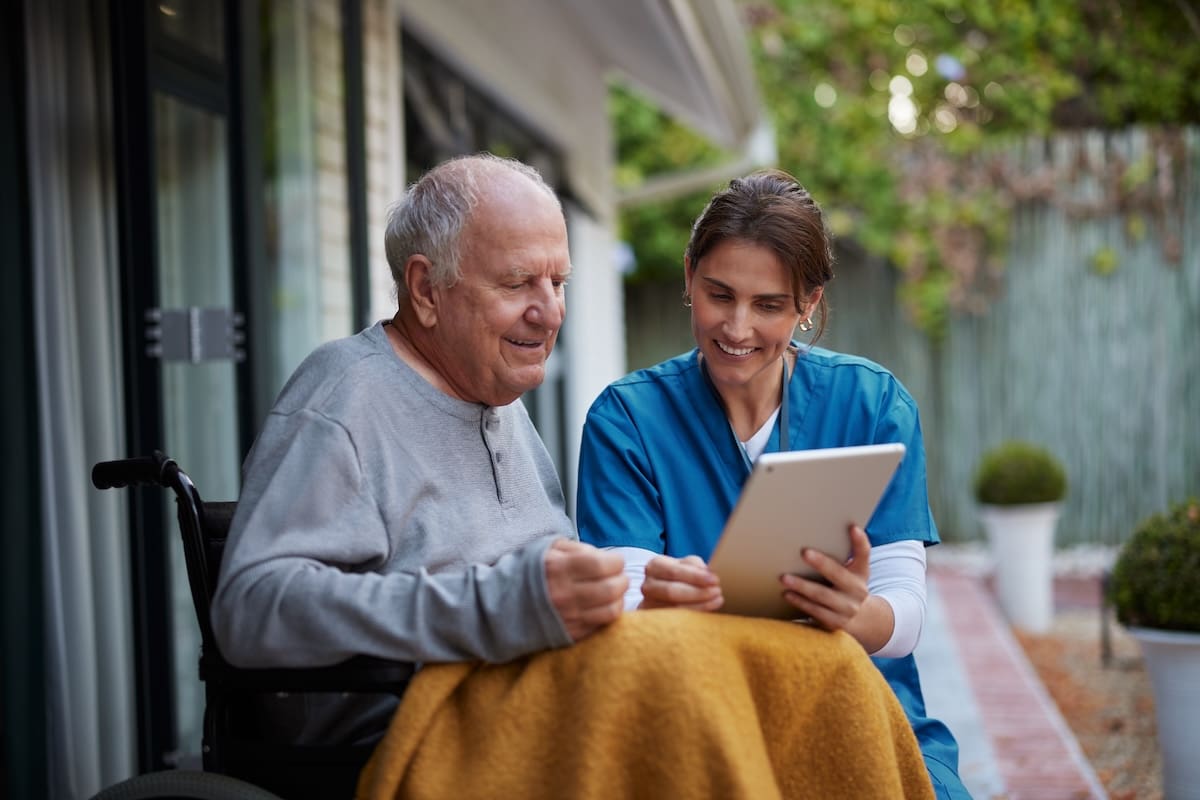 Caregiver in blue scrubs showing a tablet to an elderly man seated in a wheelchair outdoors with a blanket over his lap.