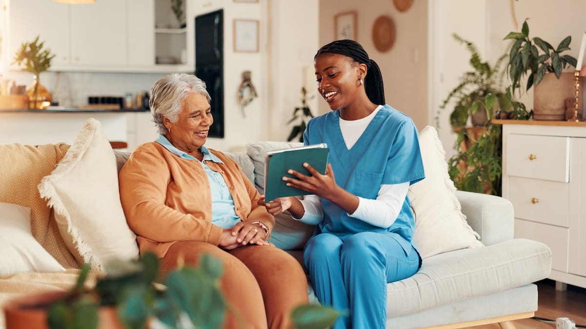 Resident and caregiver in scrubs sitting on a couch engaged in a social activity using a tablet in a comfortable living room.
