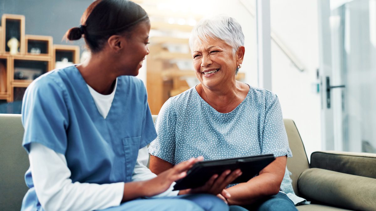 Healthcare professional engaging with a smiling senior woman using a digital tablet in a living room setting.