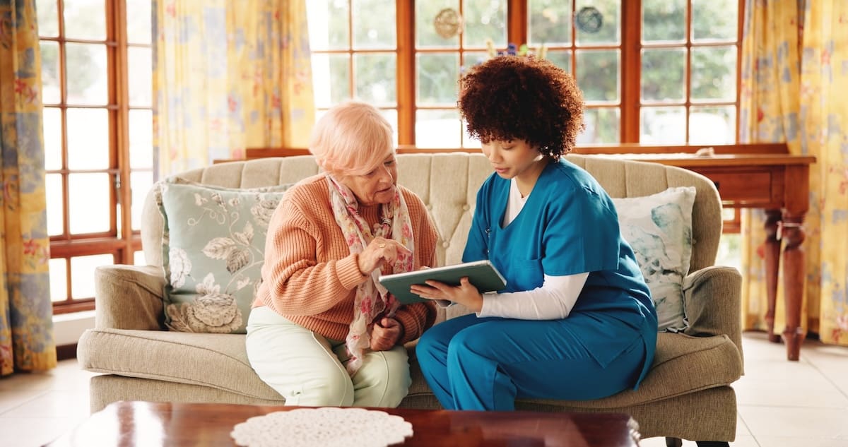 Caregiver and resident sitting on a couch together looking at a tablet in a bright living room.