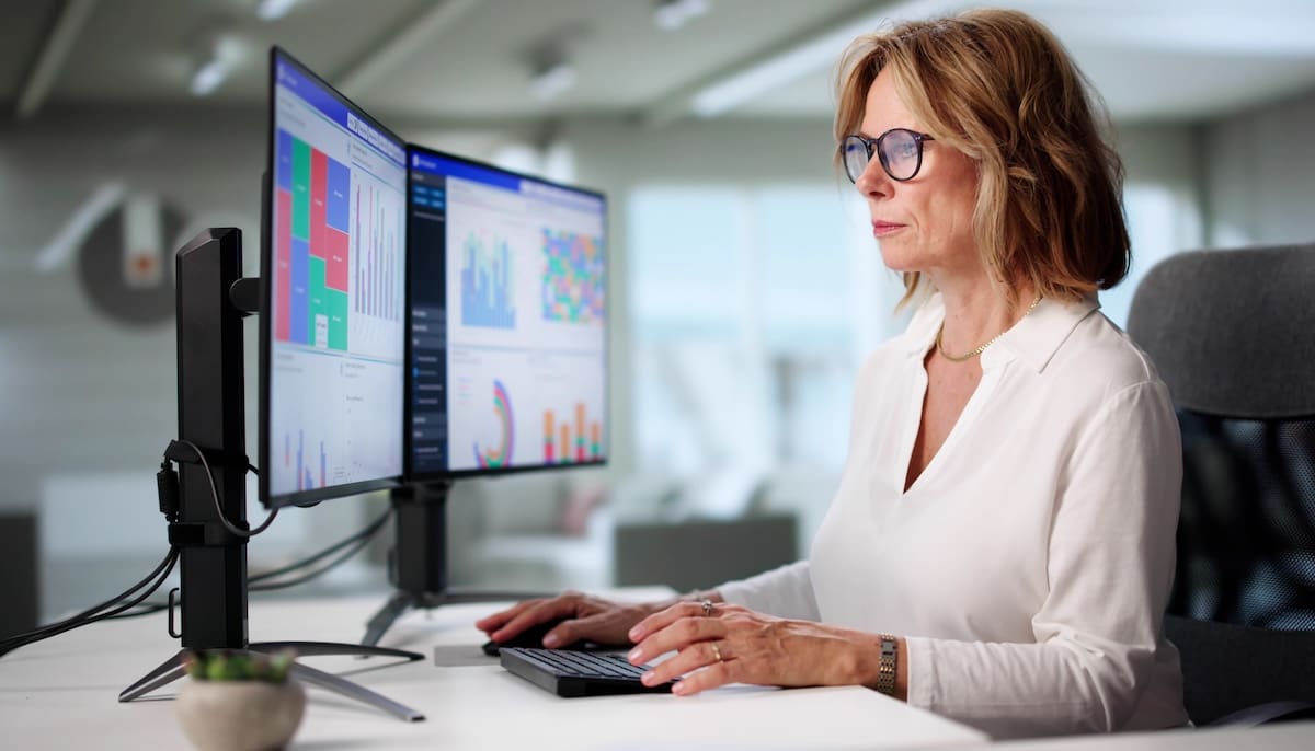 Person working at a desk with dual monitors showing charts and graphs in an office environment.