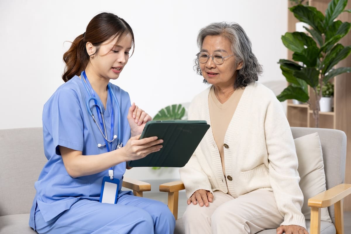 Healthcare professional in scrubs consulting with an older adult resident using a tablet while seated on a couch in a senior living care environment.