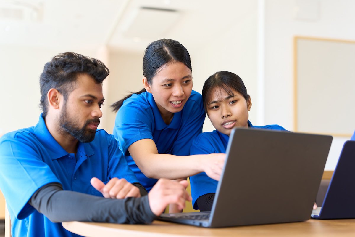 Three people seated around a table collaborating and discussing while looking at a laptop screen.