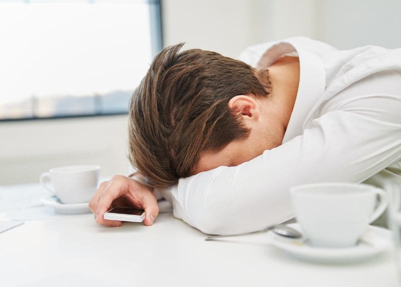 Person resting their head on a table holding a smartphone near two coffee cups indoors.
