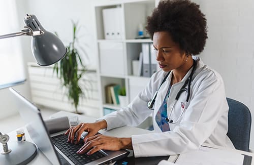 A healthcare professional typing on a laptop at a desk in a well-lit office environment with medical equipment.