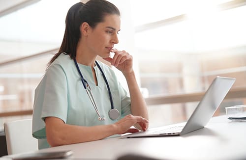 Healthcare professional in scrubs working on a laptop in a bright care environment.