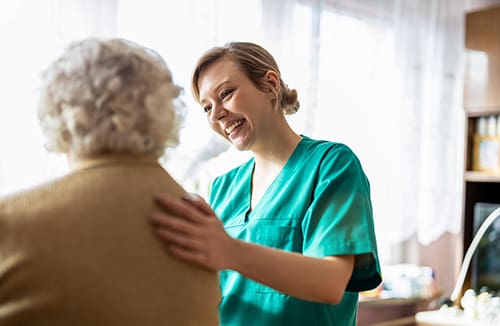 Caregiver in teal uniform gently supporting an elderly person in a brown sweater in a warmly lit room.